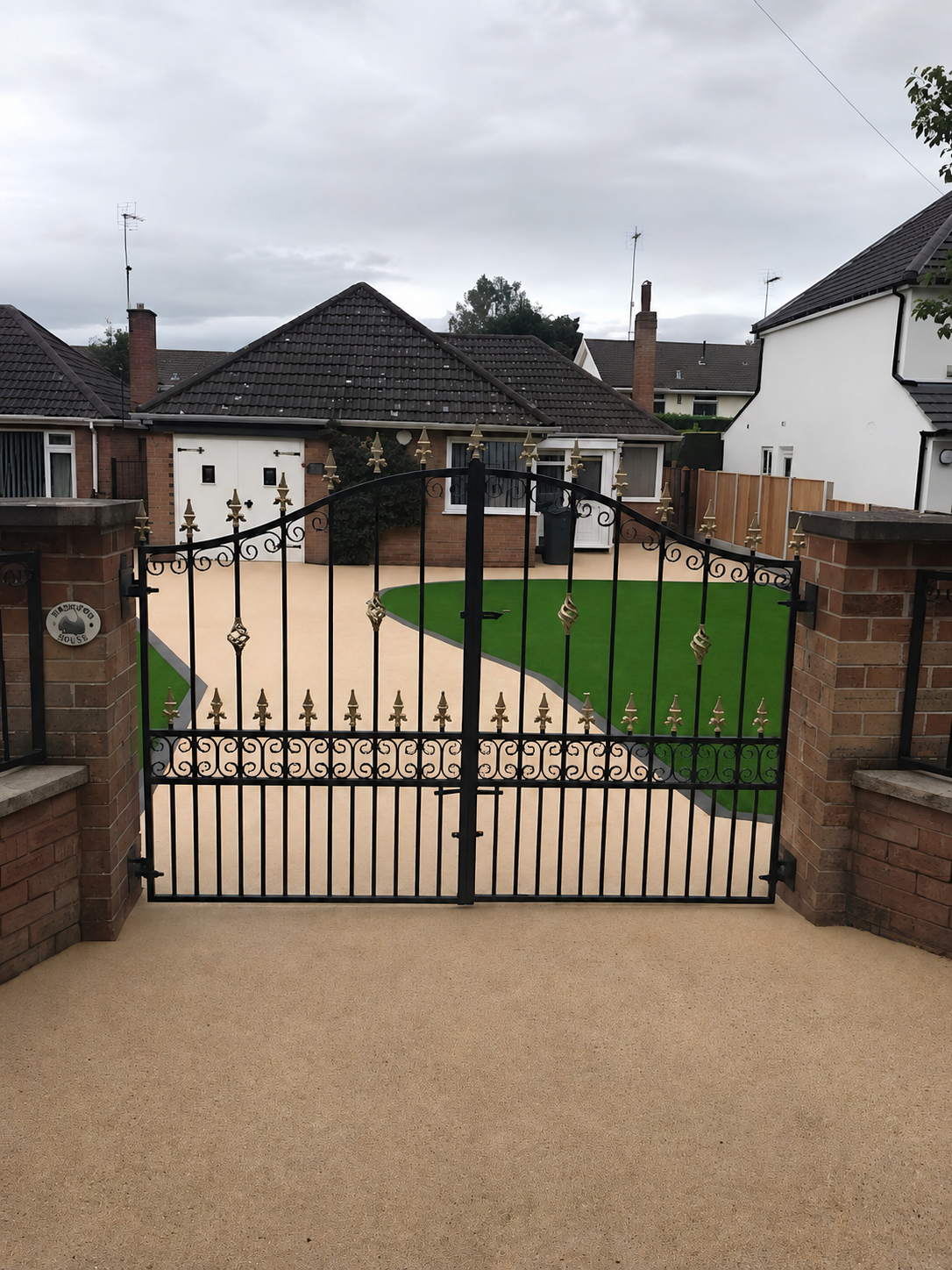 Finished resin bound driveway seen front-on through closed black wrought iron gates with artificial grass either side